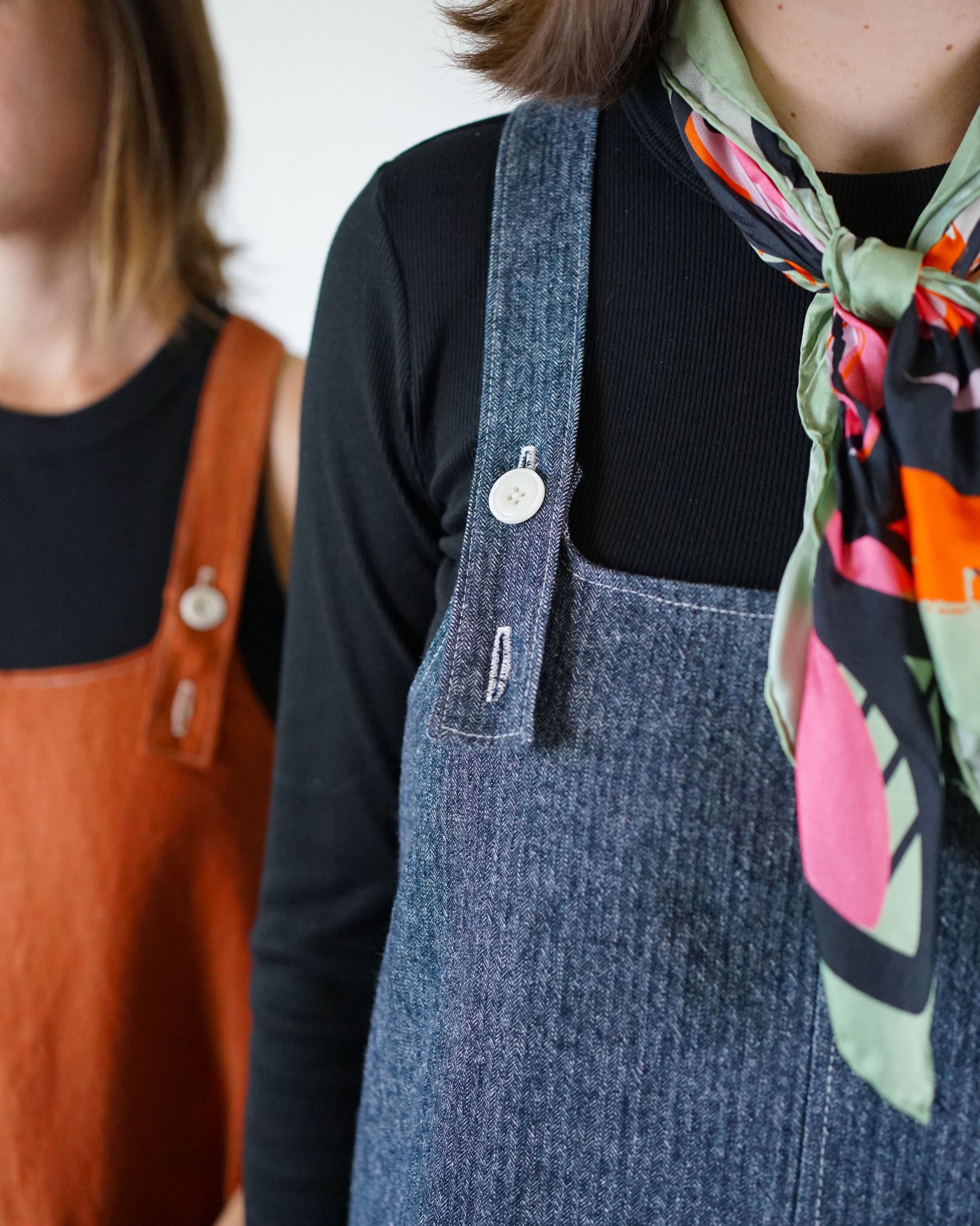 Person wearing a denim pinafore dress with a colorful scarf, standing next to another person in an orange dress.
