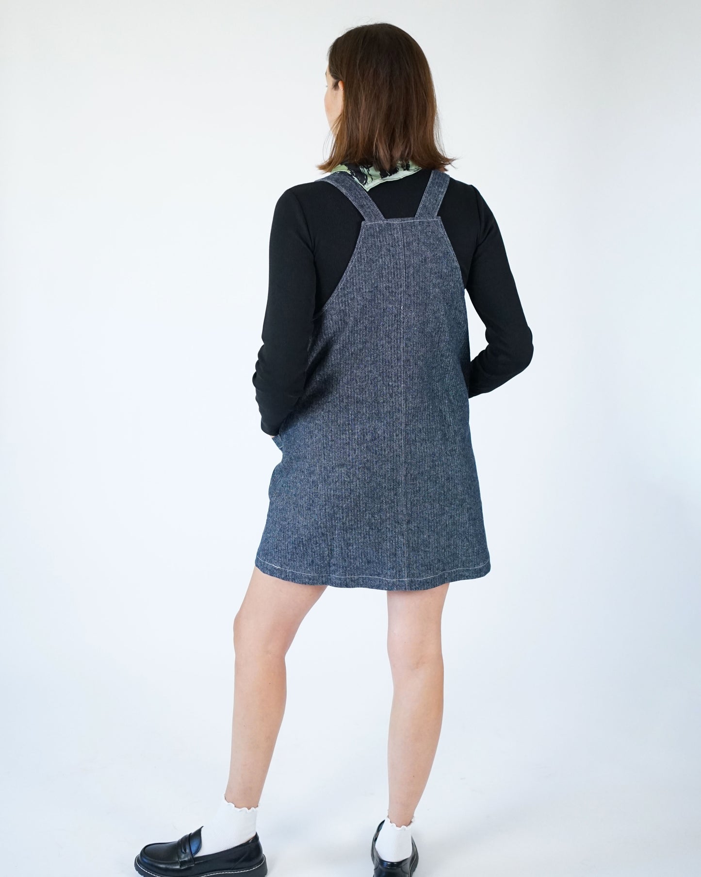 Person wearing a denim pinafore dress over a black top on a white background