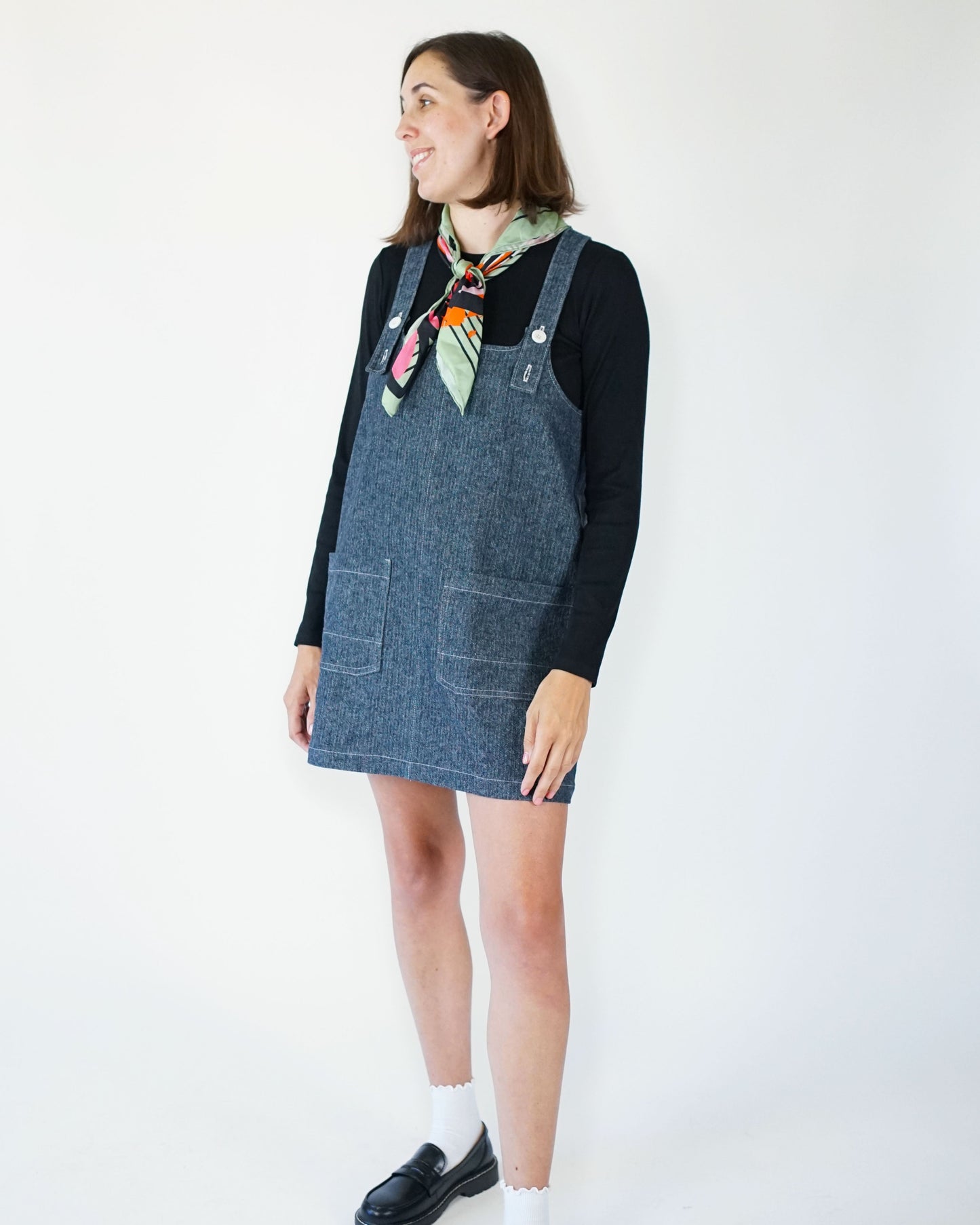 Woman wearing a denim pinafore dress with a black long-sleeve shirt and colorful scarf on a white background