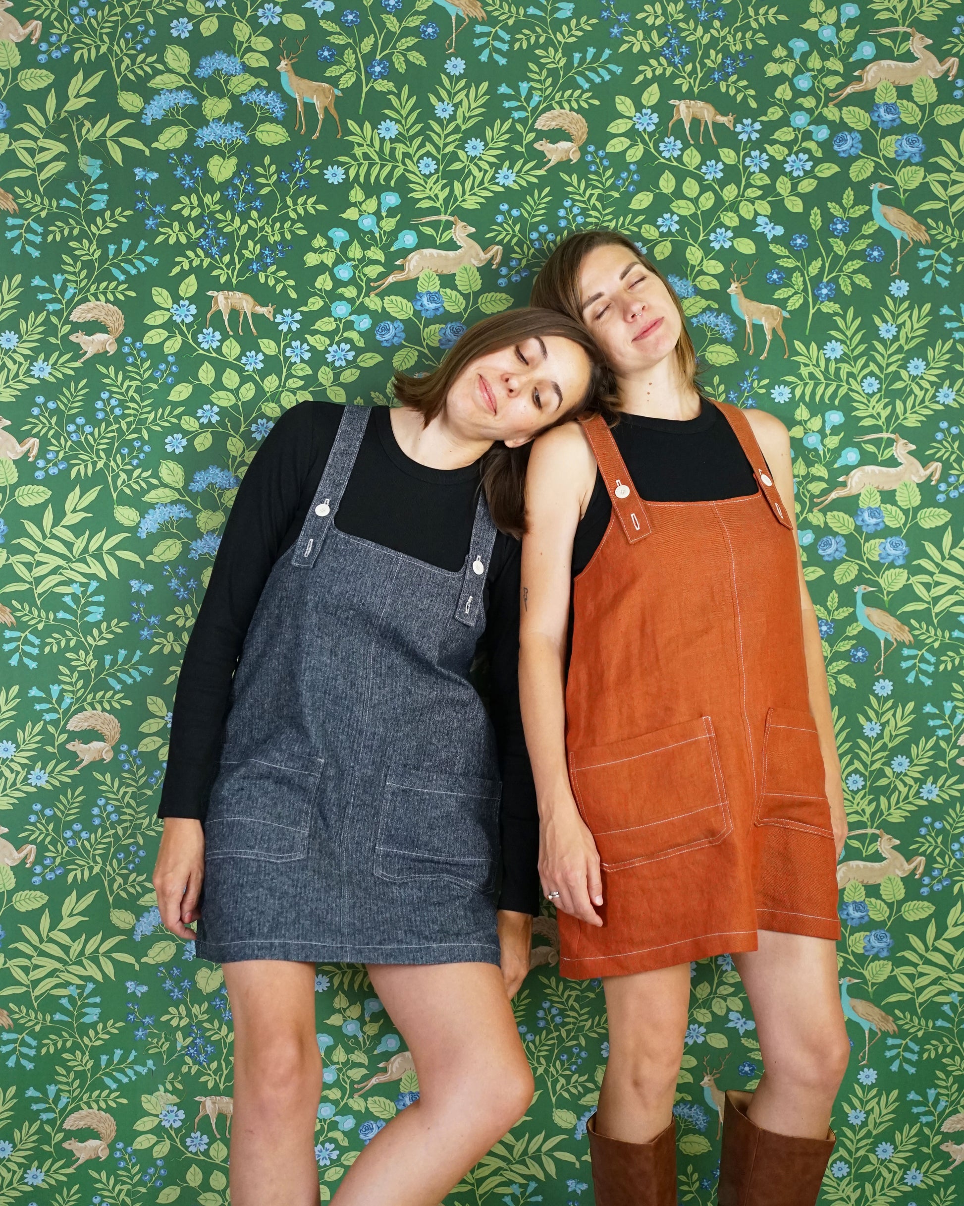 Two women wearing denim and brown pinafore dresses against a patterned wallpaper background.