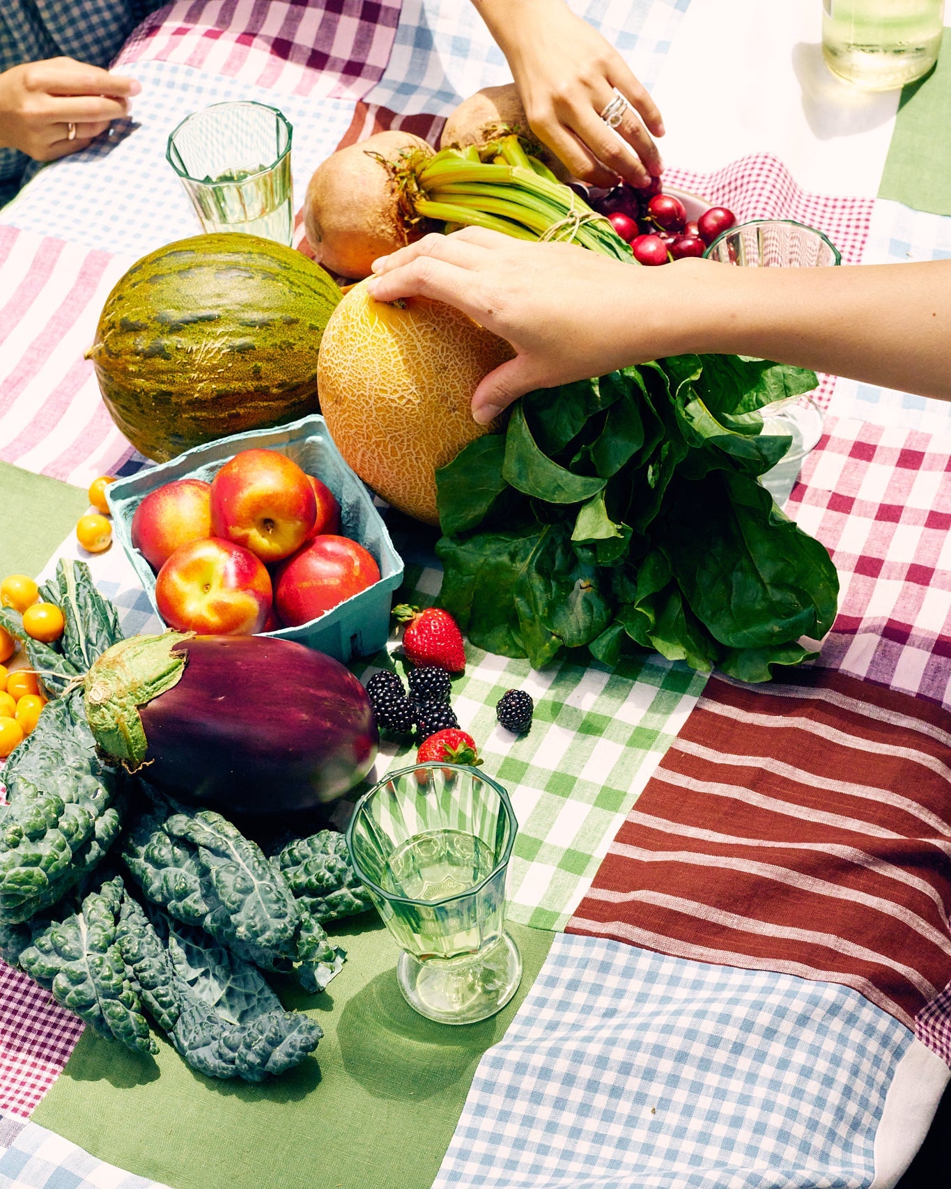 Assorted fruits and vegetables on a linen quilt with glasses of water.