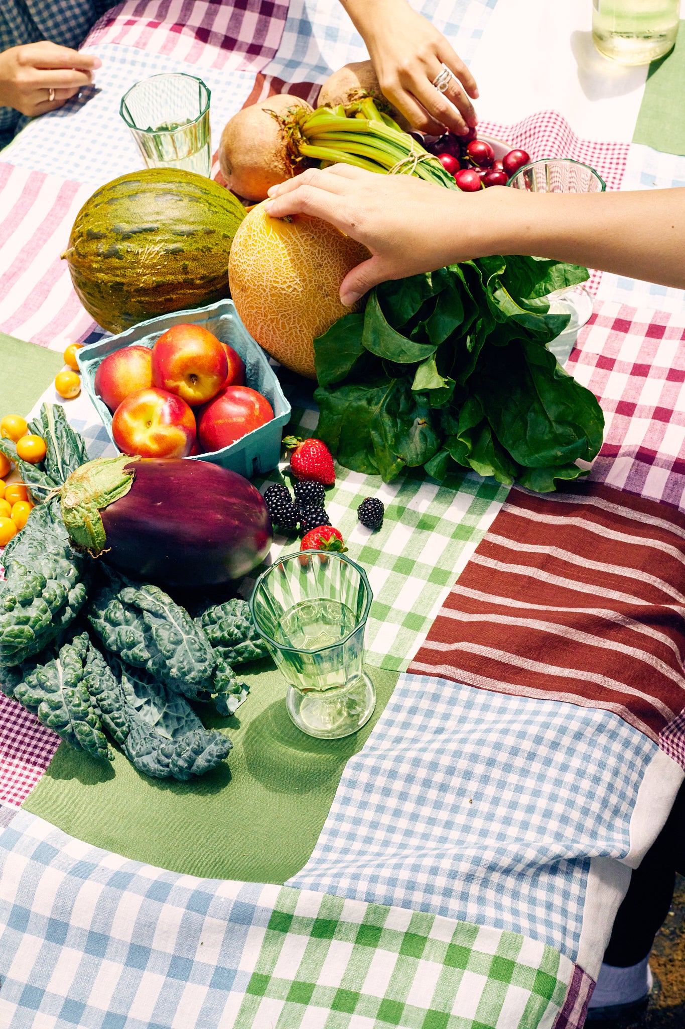 Assorted fruits and vegetables on a linen quilt with glasses of water.