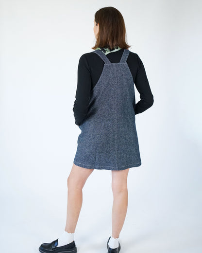 Person wearing a denim pinafore dress over a black top on a white background
