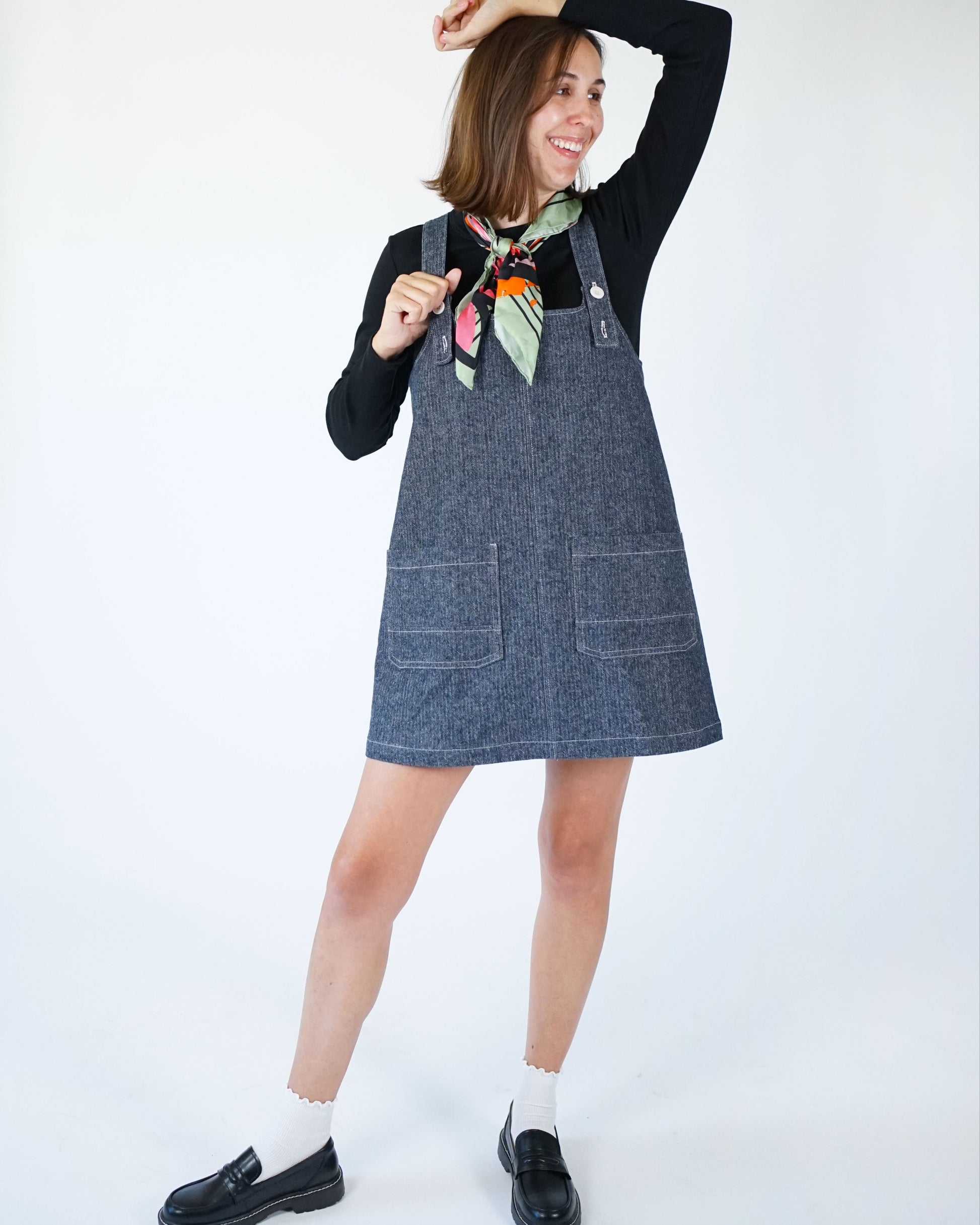 Woman wearing a denim pinafore dress with a black top against a white background