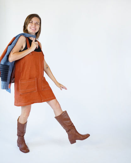 Woman wearing an orange dress and brown boots with a blue sweater over her shoulder on a white background